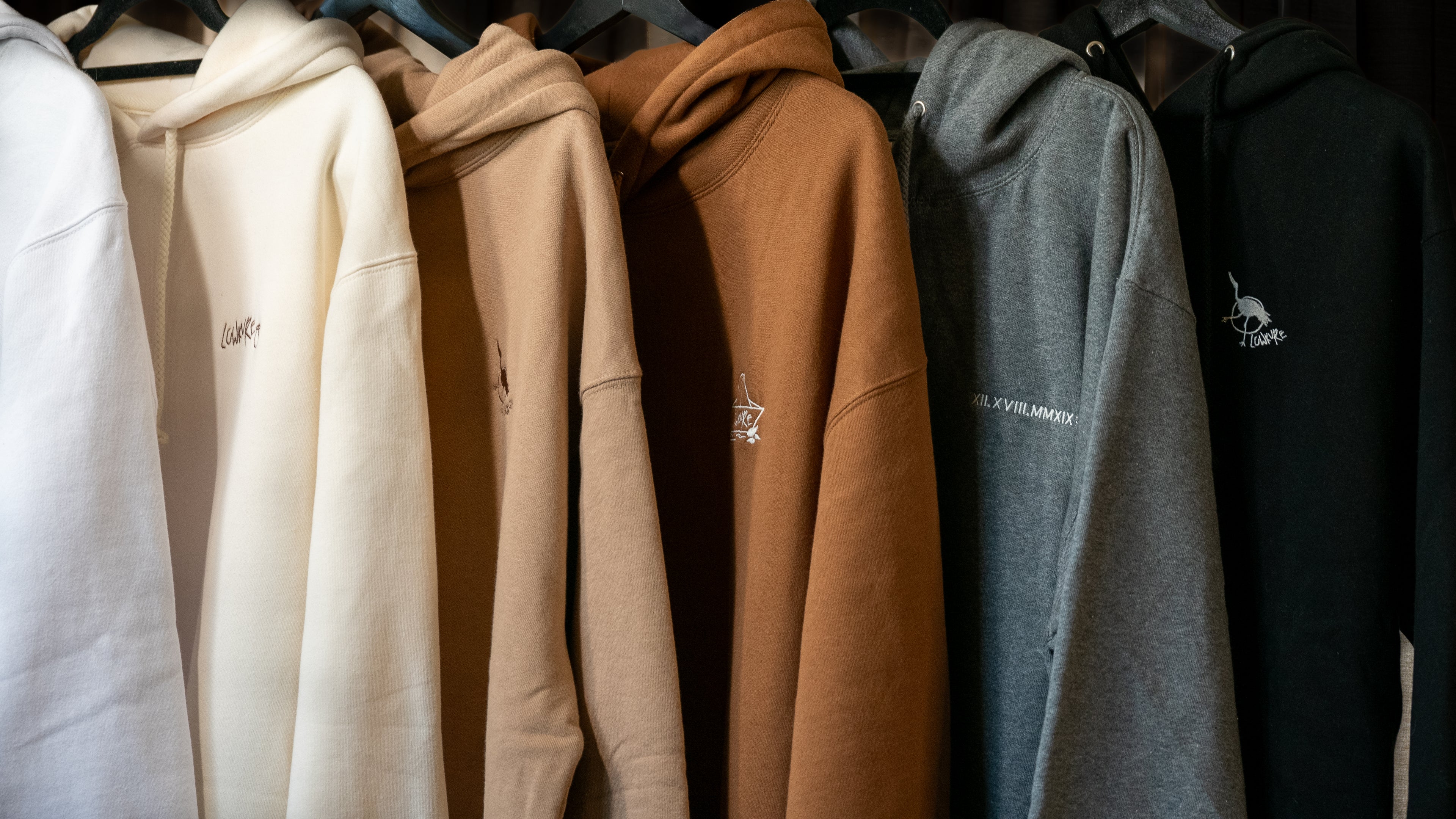 Row of LowKure hoodies in various colours - white, bone, sandstorm, saddle, heather grey, black - showcasing each LowKure Logo. LowKure TearLight, a symbol of healing. River of Tears - A symbol of resilience. The Japanese crane, a symbol of hope. A Sister's Promise, a symbol of love.